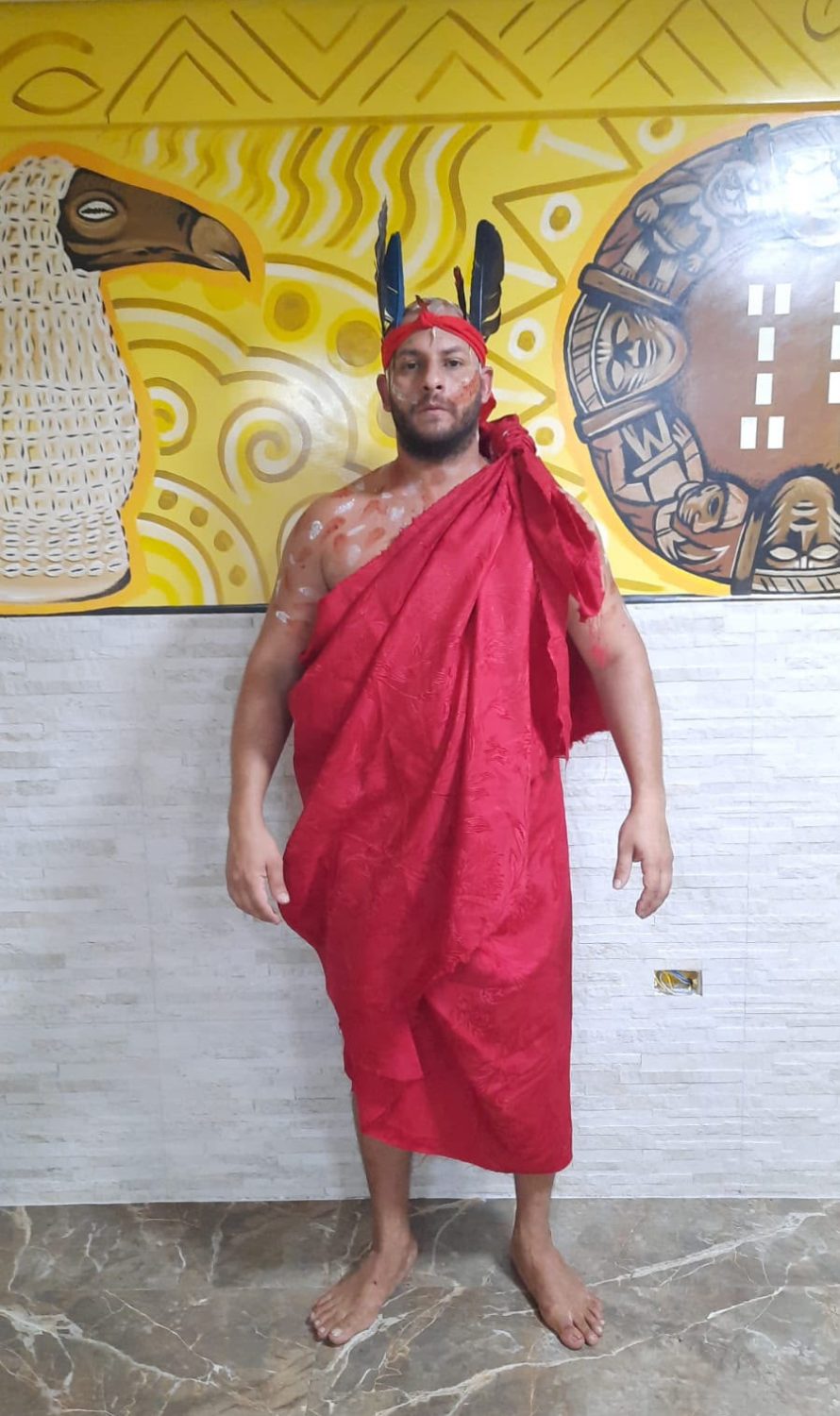 Dirigente do Templo da Serpente Negra em manto ritual vermelho com cocar diante de painel artístico